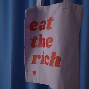 Puede incluir: Una bolsa de tela de color morado claro con el texto rojo "eat the rich" impreso en una fuente negrita y redondeada en la parte frontal. La bolsa incluye dos correas de hombro de tela a juego y un peque&ntilde;o punto rojo bajo el texto.