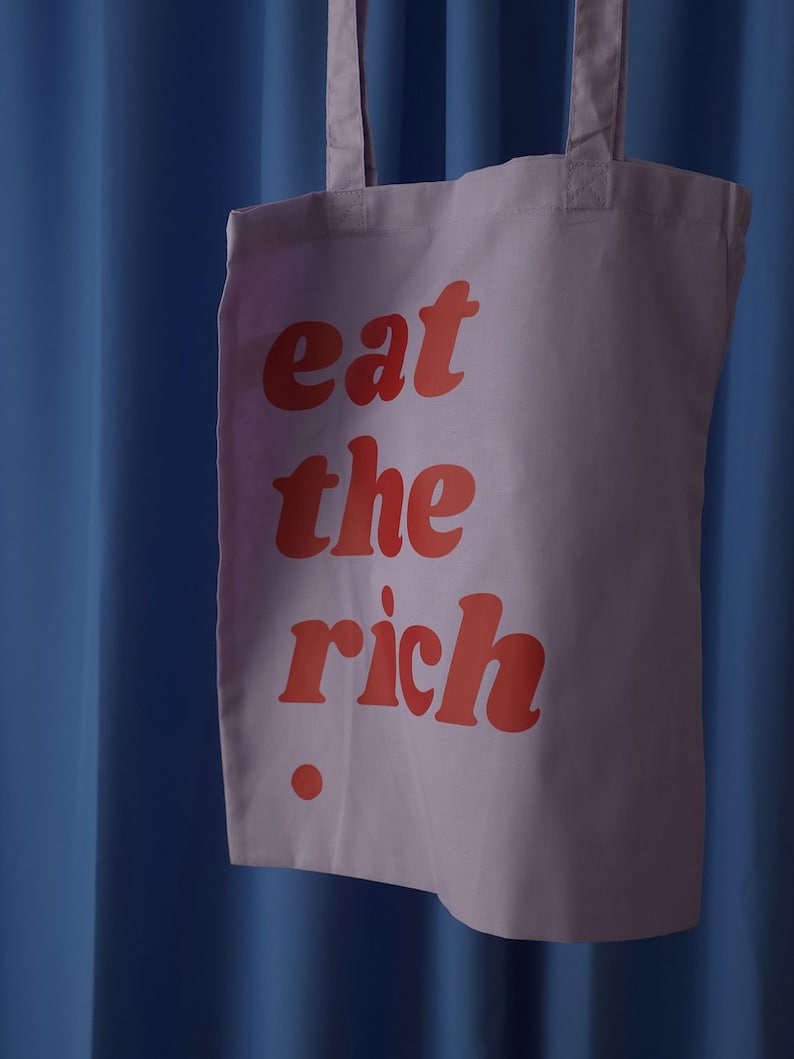 Puede incluir: Una bolsa de tela de color morado claro con el texto rojo "eat the rich" impreso en una fuente negrita y redondeada en la parte frontal. La bolsa incluye dos correas de hombro de tela a juego y un peque&ntilde;o punto rojo bajo el texto.