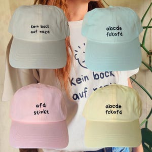 Op de afbeelding: Vier verschillende baseballpetten met verschillende slogans geborduurd erop. De petten zijn in verschillende kleuren: lichtblauw, lichtroze, lichtgeel en wit. De slogans zijn: "kein bock auf nazis", "afD stinkt", "abcde fckafd" en "abcde fckafd".