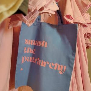 Puede incluir: Una bolsa de tela azul claro con dos asas que presenta el texto rosa "smash the patriarchy" impreso en la parte frontal. La bolsa cuelga frente a una pila de camisetas de color rosa claro dobladas, mostrando un contraste de color evidente.