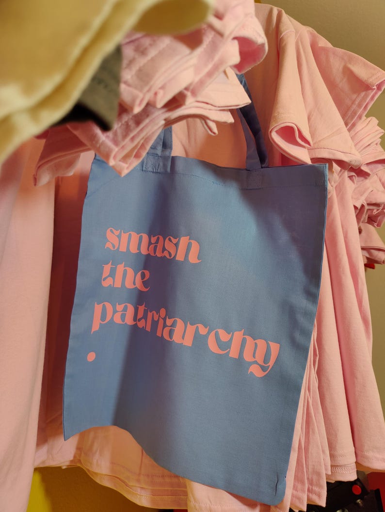 Puede incluir: Una bolsa de tela azul claro con dos asas que presenta el texto rosa "smash the patriarchy" impreso en la parte frontal. La bolsa cuelga frente a una pila de camisetas de color rosa claro dobladas, mostrando un contraste de color evidente.