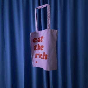 Könnte beinhalten: Eine hellviolette Tragetasche mit zwei Schulterriemen, die auf der Vorderseite den roten Schriftzug "eat the rich" in einer fetten, abgerundeten Schriftart zeigt, ergänzt durch einen kleinen roten Punkt unter dem Wort "rich" unten links.