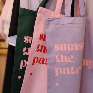 Puede incluir: Tres bolsas de tela rectangulares con dos asas cada una, colgadas en fila. Las bolsas son de color verde oscuro, rosa claro y morado claro. Cada bolsa presenta el texto "smash the patriarchy" impreso en una fuente negrita y estilizada.