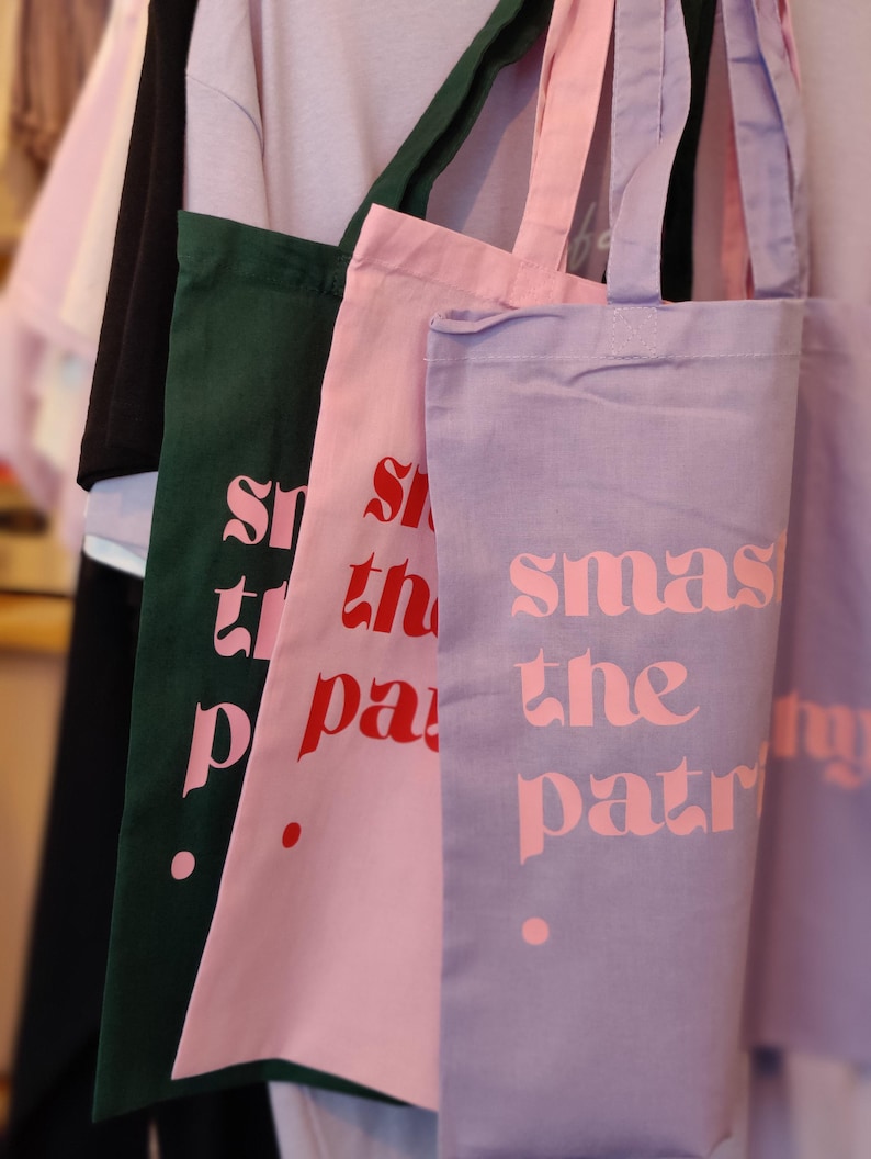 Puede incluir: Tres bolsas de tela rectangulares con dos asas cada una, colgadas en fila. Las bolsas son de color verde oscuro, rosa claro y morado claro. Cada bolsa presenta el texto "smash the patriarchy" impreso en una fuente negrita y estilizada.