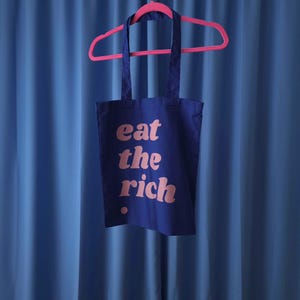 Op de afbeelding: Een donkerblauwe draagtas hangend aan een felroze plastic kledinghanger. De tas is aan de voorzijde bedrukt met de tekst "eat the rich" in een lichtroze, afgerond lettertype, vergezeld van een kleine roze stip onder de tekst.