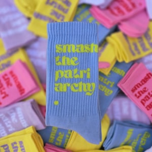 May include: A light blue ribbed crew sock featuring the text "smash the patriarchy" in a bright yellow font, with a small yellow dot positioned below the text. The sock is displayed against a background of various other colorful socks.