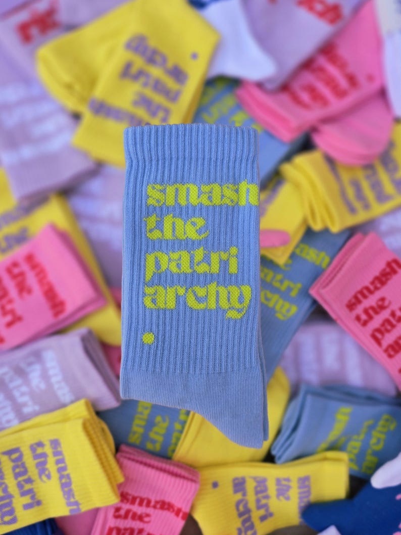 May include: A light blue ribbed crew sock featuring the text "smash the patriarchy" in a bright yellow font, with a small yellow dot positioned below the text. The sock is displayed against a background of various other colorful socks.