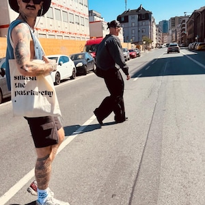 Puede incluir: Un bolso de tela color crema con el texto impreso en negro 'smash the patriarchy' y un peque&ntilde;o punto al final. El bolso lo lleva al hombro una persona que viste un chaleco de punto azul, pantalones cortos negros, un sombrero y zapatillas.