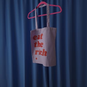 Puede incluir: Una bolsa de tela de color morado claro colgada de una percha de pl&aacute;stico de color rosa brillante. La bolsa presenta el texto "eat the rich" impreso en una fuente roja, negrita y redondeada en la parte frontal, con un peque&ntilde;o punto rojo.