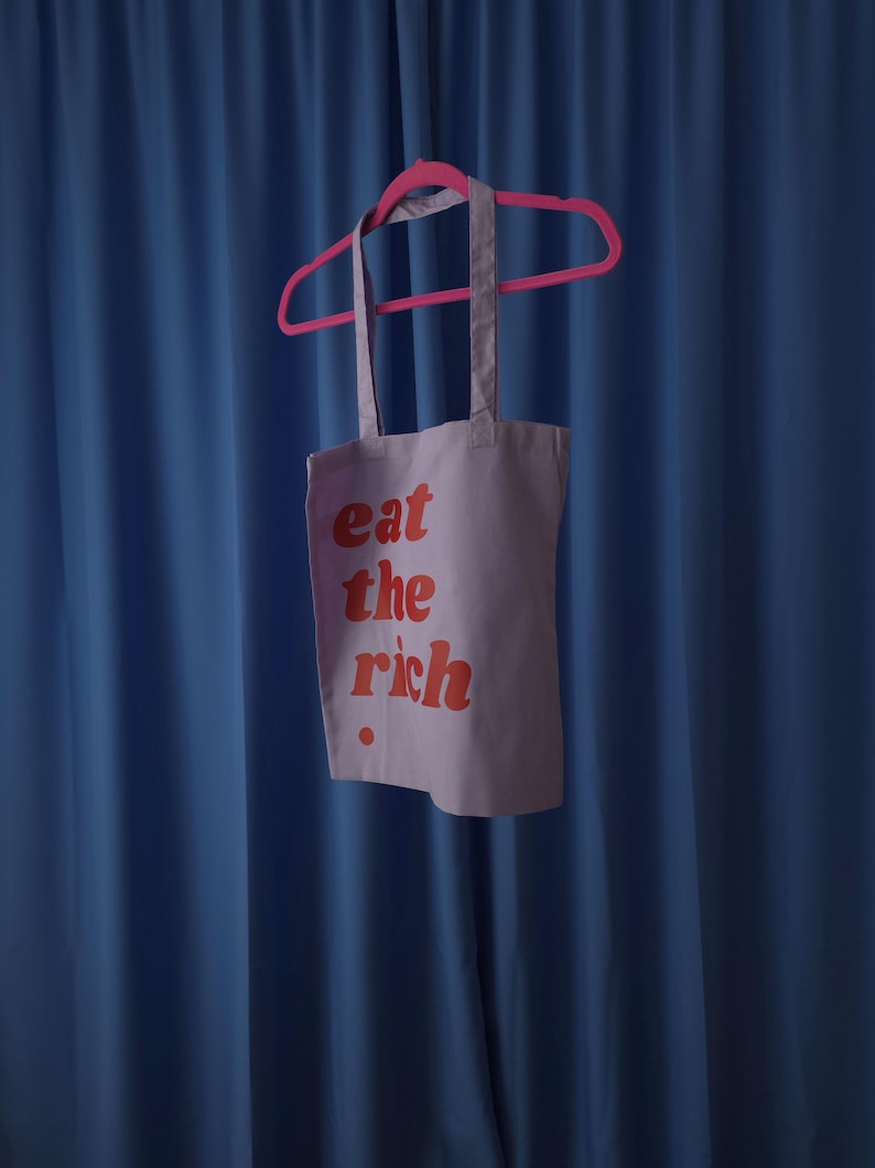 Puede incluir: Una bolsa de tela de color morado claro colgada de una percha de pl&aacute;stico de color rosa brillante. La bolsa presenta el texto "eat the rich" impreso en una fuente roja, negrita y redondeada en la parte frontal, con un peque&ntilde;o punto rojo.