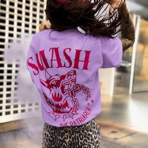 Könnte beinhalten: Ein hellviolettes T-Shirt mit einer dunkelrosa Grafik eines knurrenden Hundes, der eine Kette sprengt, sowie dem dunkelrosa Schriftzug SMASH THE PATRIARCH. Dazu werden eine rote Baseballkappe und ein Rock mit Leopardenmuster getragen.