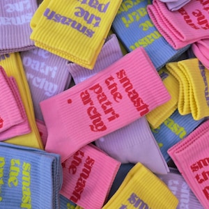 May include: A collection of ribbed crew socks in various pastel colors including yellow, pink, light blue, and lavender. Each sock features the text "smash the patriarchy." printed in a contrasting color on the side of the ribbed fabric.