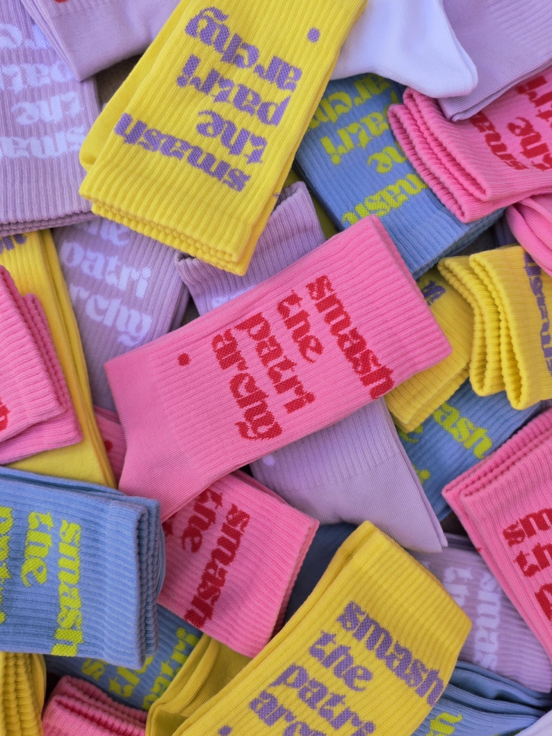 May include: A collection of ribbed crew socks in various pastel colors including yellow, pink, light blue, and lavender. Each sock features the text "smash the patriarchy." printed in a contrasting color on the side of the ribbed fabric.