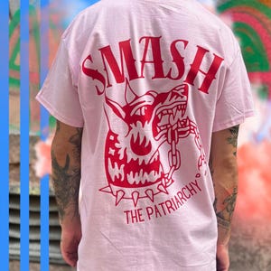 Könnte beinhalten: Hellrosa T-Shirt mit roter Grafik auf dem Rücken. Die Grafik zeigt das Wort "SMASH" über einer grimmigen Hundeillustration mit dem Text "THE PATRIARCHY" darunter. Das T-Shirt hat kurze Ärmel.