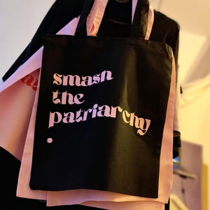 Puede incluir: Una bolsa de tela negra con dos asas que presenta el texto "smash the patriarchy" impreso en un degradado de letras rosas y naranjas. La bolsa se muestra sobre un fondo de tela rosa claro, exhibiendo un dise&ntilde;o tipogr&aacute;fico claro y llamativo.