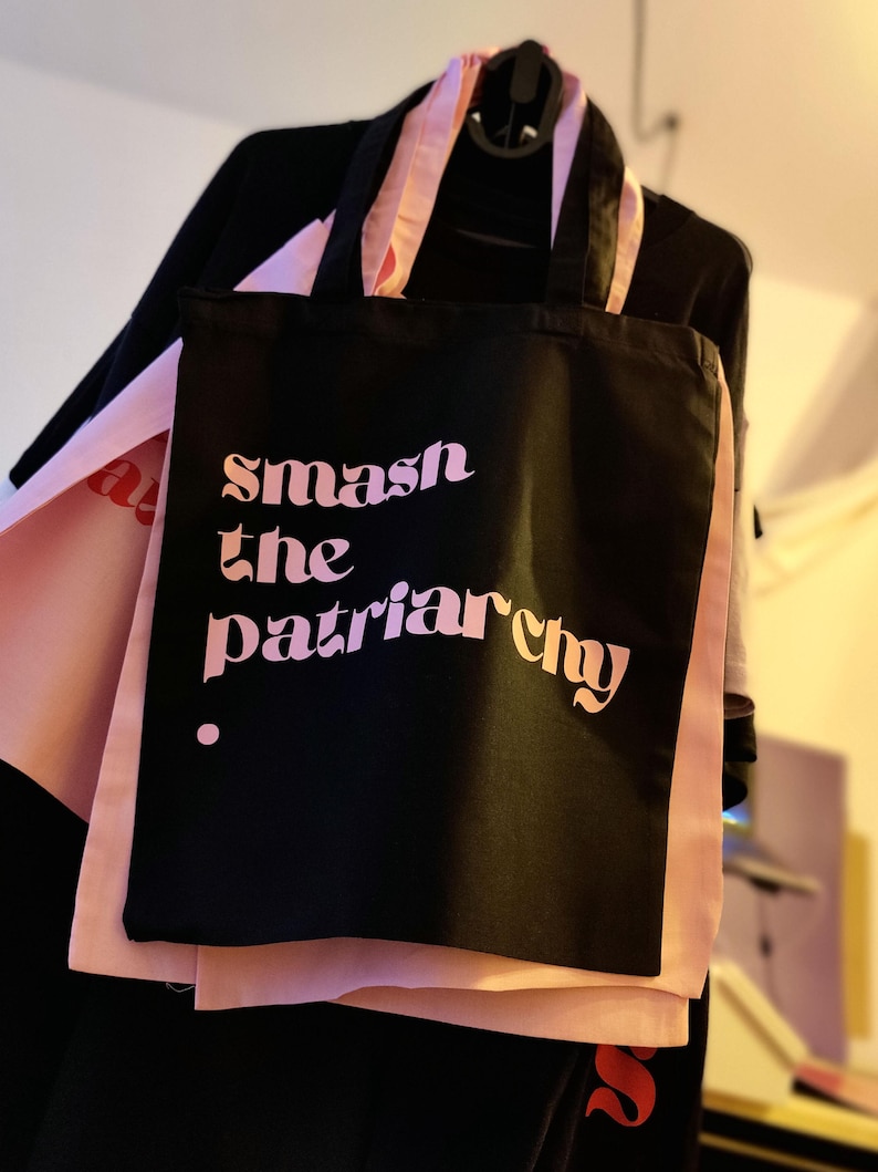 Puede incluir: Una bolsa de tela negra con dos asas que presenta el texto "smash the patriarchy" impreso en un degradado de letras rosas y naranjas. La bolsa se muestra sobre un fondo de tela rosa claro, exhibiendo un dise&ntilde;o tipogr&aacute;fico claro y llamativo.