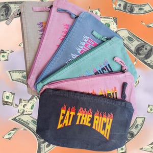 Könnte beinhalten: Eine Sammlung von Reißverschluss-Federmäppchen in verschiedenen Farben, darunter Grau, Rosa, Blau und Türkis. Jedes Etui zeigt den Schriftzug "EAT THE RICH" in Gelb mit einem Flammendesign. Im Hintergrund fallen Geldscheine.