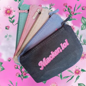 Könnte beinhalten: Eine Auswahl an Denim-Reißverschlusstaschen in verschiedenen Farben: Türkis, Rosa, Beige, Hellblau und Dunkelgrau. Die dunkelgraue Tasche trägt den rosa Schriftzug "Macker, lol". Auf rosa floralem Hintergrund.