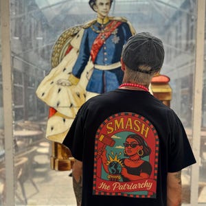Può includere: T-shirt nero con grafica "SMASH The Patriarchy". La grafica presenta una donna che tiene un martello sopra un globo. Sullo sfondo, un grande dipinto di un uomo in uniforme blu e oro.