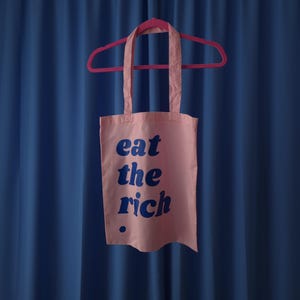 Puede incluir: Una bolsa de tela rosa claro colgada de una percha rosa brillante. La bolsa presenta el texto "eat the rich" impreso en una fuente redondeada, negrita y azul oscuro, con un peque&ntilde;o punto azul oscuro debajo del texto en la parte inferior.