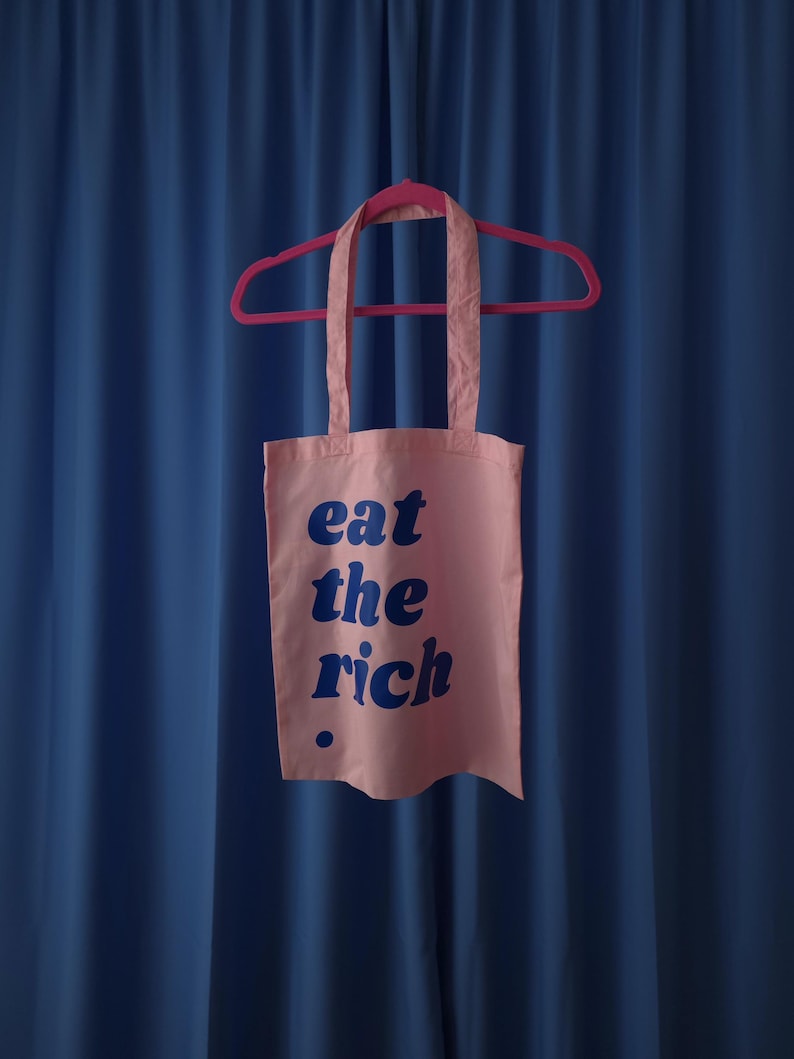 Puede incluir: Una bolsa de tela rosa claro colgada de una percha rosa brillante. La bolsa presenta el texto "eat the rich" impreso en una fuente redondeada, negrita y azul oscuro, con un peque&ntilde;o punto azul oscuro debajo del texto en la parte inferior.