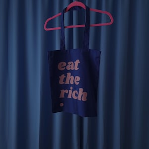 Puede incluir: Una bolsa de tela de algod&oacute;n azul oscuro colgada de una percha de pl&aacute;stico rosa brillante. La bolsa presenta el texto "eat the rich" impreso en una fuente redondeada de color rosa claro en el frente, con un peque&ntilde;o punto rosa debajo.