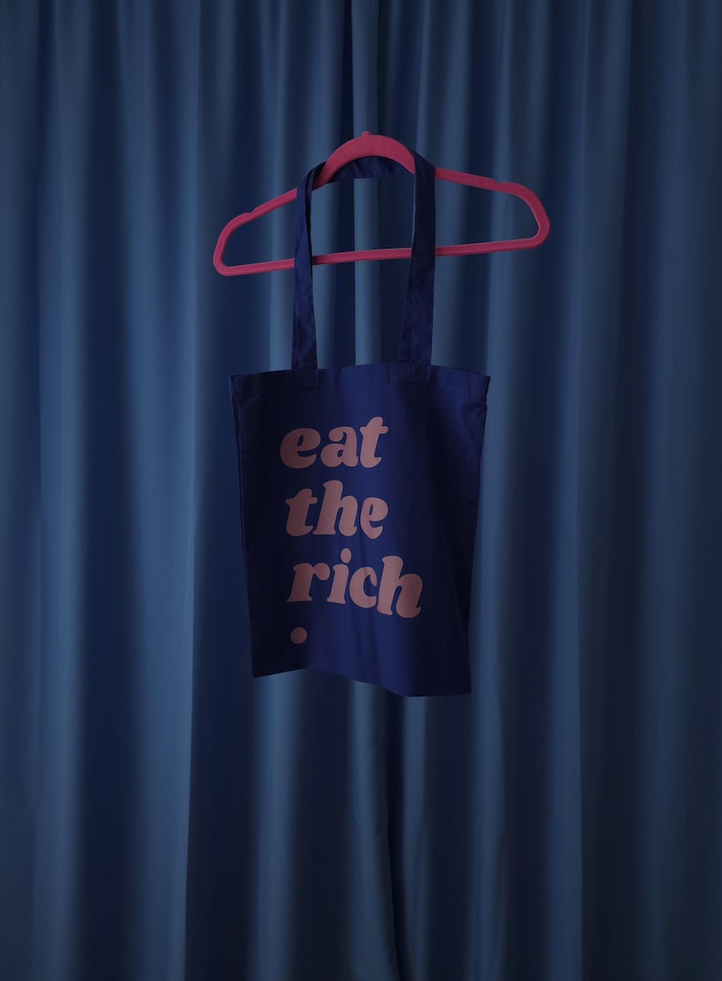 Puede incluir: Una bolsa de tela de algod&oacute;n azul oscuro colgada de una percha de pl&aacute;stico rosa brillante. La bolsa presenta el texto "eat the rich" impreso en una fuente redondeada de color rosa claro en el frente, con un peque&ntilde;o punto rosa debajo.
