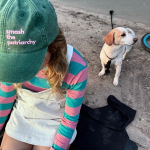 Könnte beinhalten: Eine Person trägt eine grüne Baseballkappe mit dem Text "smash the patriarchy" in rosa Buchstaben, ein pink-türkisfarbenes, langärmliges Hemd und ein weißes Latzkleid. Die Person sitzt auf einem türkisfarbenen Skateboard mit einem schwarz-weißen Aufkleber. Ein weiß-brauner Hund sitzt neben der Person auf dem Boden.