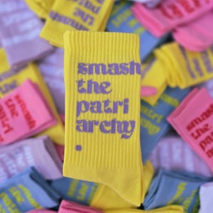 May include: A bright yellow ribbed crew sock featuring the purple text "smash the patriarchy" printed vertically in a bold, sans-serif font, accompanied by a small purple dot at the bottom left of the text.