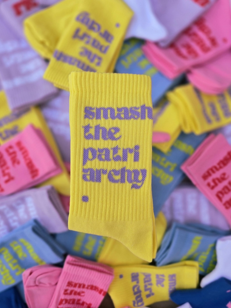 May include: A bright yellow ribbed crew sock featuring the purple text "smash the patriarchy" printed vertically in a bold, sans-serif font, accompanied by a small purple dot at the bottom left of the text.