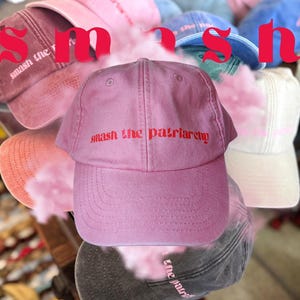 Puede incluir: Una pila de gorras de béisbol rosas, azules y blancas con la frase "smash the patriarchy" en letras rojas en la parte delantera de una gorra rosa.