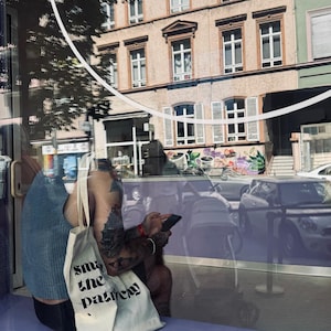 Puede incluir: Una camiseta sin mangas de punto azul claro y una bolsa de tela color crema con el texto impreso en negro "smash the patriarchy". La bolsa cuenta con dos correas para el hombro y est&aacute; hecha de tela tejida, vista a nivel de la calle.