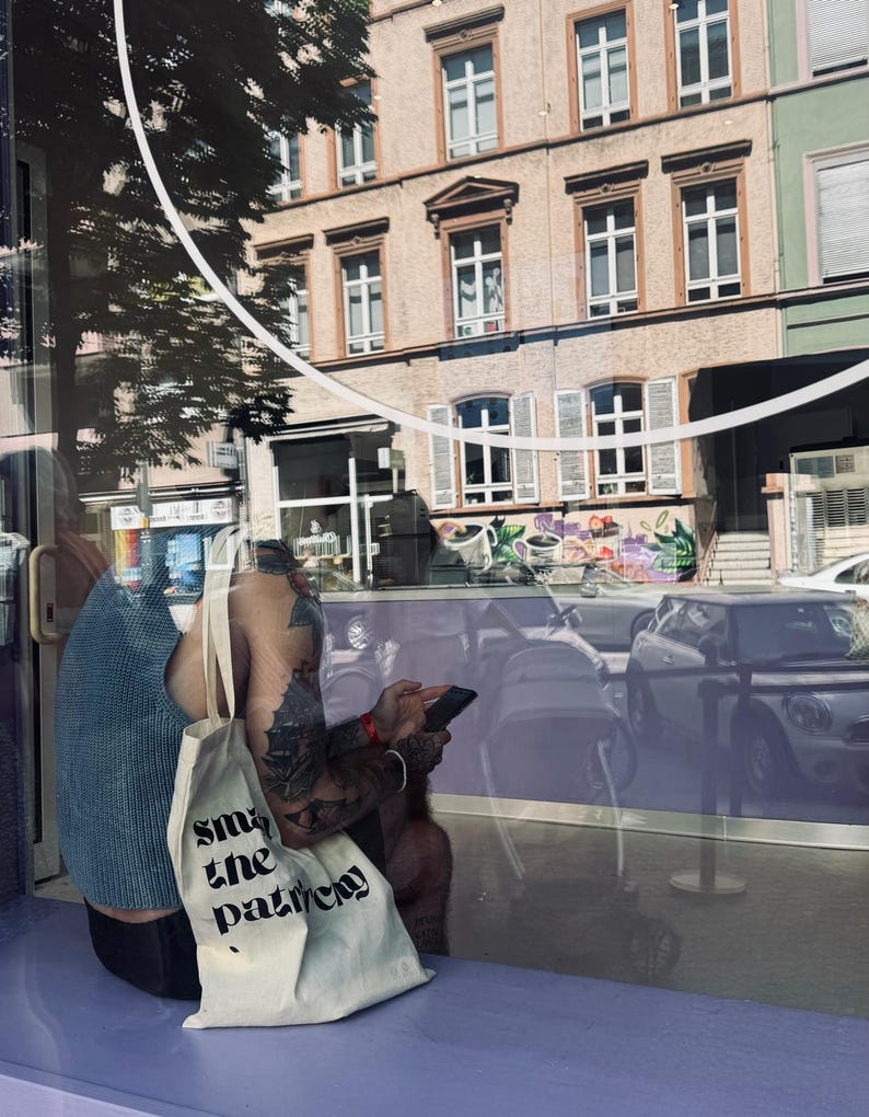 Puede incluir: Una camiseta sin mangas de punto azul claro y una bolsa de tela color crema con el texto impreso en negro "smash the patriarchy". La bolsa cuenta con dos correas para el hombro y est&aacute; hecha de tela tejida, vista a nivel de la calle.
