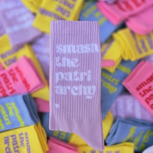 May include: A light purple ribbed crew sock featuring the white text "smash the patriarchy" printed vertically on the side, with a small white dot below the text. The sock is positioned in front of a pile of similar socks in various colors.