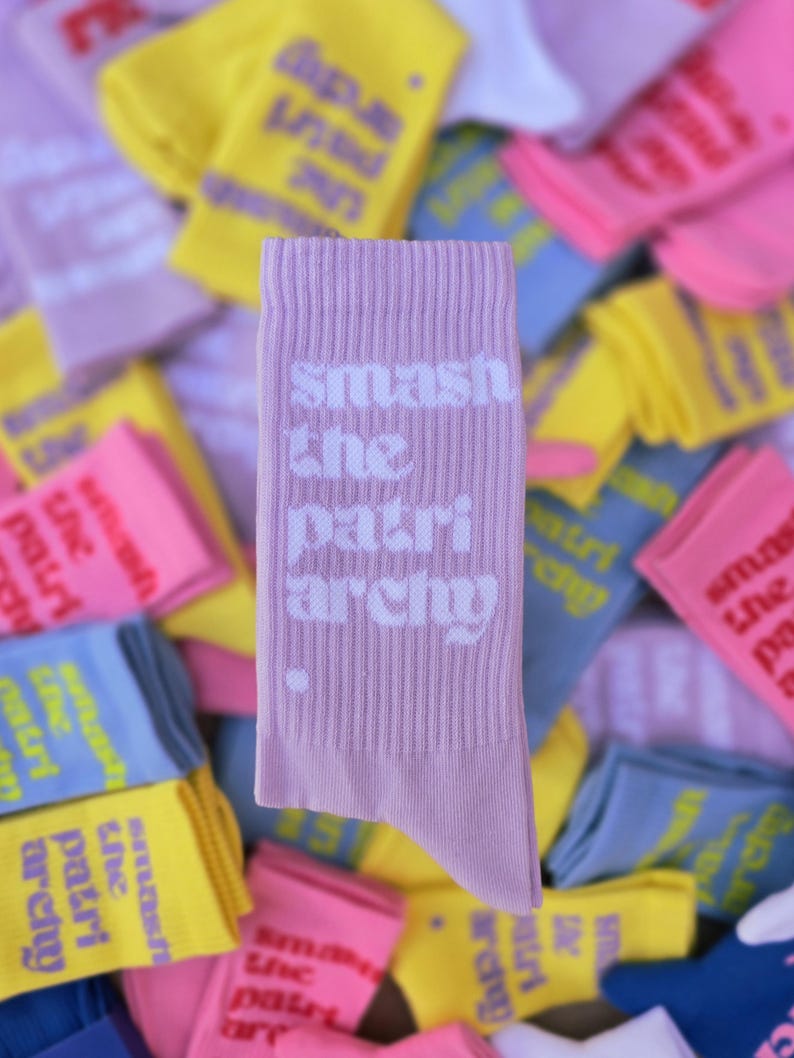 May include: A light purple ribbed crew sock featuring the white text "smash the patriarchy" printed vertically on the side, with a small white dot below the text. The sock is positioned in front of a pile of similar socks in various colors.