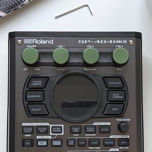 Può includere: Primo piano di un campionatore Roland SP-404MKII grigio scuro con manopole verdi. Il campionatore ha un display centrale e una chiave esagonale argentata sopra. Per la produzione musicale.