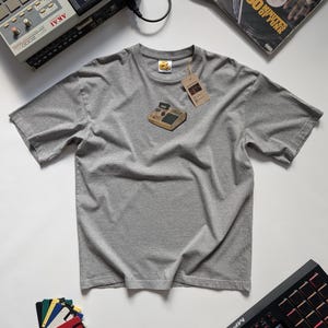 Puede incluir: Una camiseta gris jaspeado con un gráfico de un sampler vintage. La camiseta tiene una etiqueta adjunta. Otros equipos de producción musical, incluyendo un sampler y discos de vinilo, son visibles en el fondo.