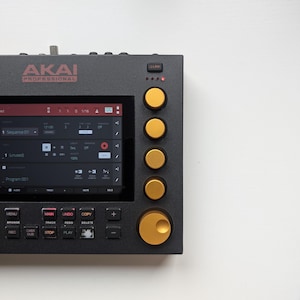 May include: A black AKAI Professional music production device with a screen displaying a user interface. It has gold-colored knobs and buttons. The screen shows text and icons related to music sequencing and audio control.
