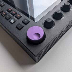 Può includere: Primo piano di un dispositivo nero per la produzione di musica elettronica con un ampio schermo e vari pulsanti e manopole. Una prominente manopola viola è visibile in basso a destra. Il dispositivo ha un design elegante e moderno.