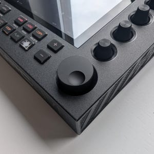 May include: Close-up of a dark gray electronic music production device. The device has a screen, buttons, and knobs. The buttons are labeled with functions like "Main", "Undo", and "Copy". The knobs are black and cylindrical.