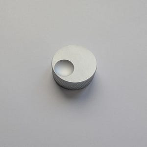 Peut inclure: Un petit bouton circulaire argent&eacute; avec un bord textur&eacute; et une indentation incurv&eacute;e en retrait. Le bouton est en m&eacute;tal et repose sur un fond uni gris clair. Le design est minimaliste et moderne.