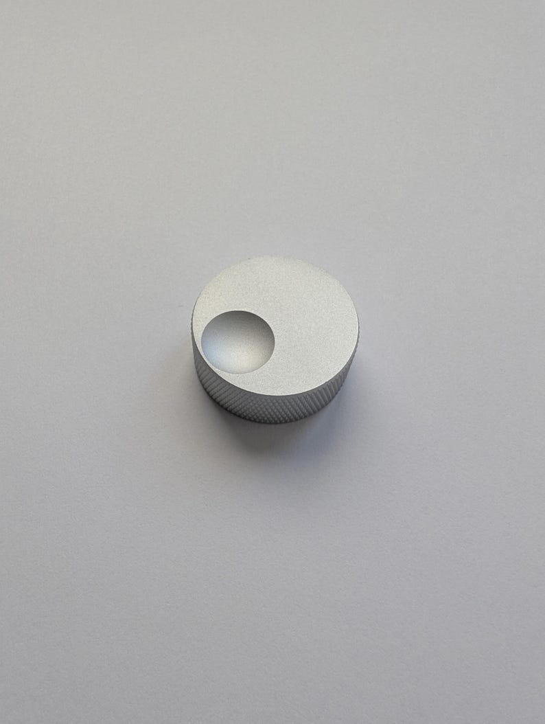 Peut inclure: Un petit bouton circulaire argent&eacute; avec un bord textur&eacute; et une indentation incurv&eacute;e en retrait. Le bouton est en m&eacute;tal et repose sur un fond uni gris clair. Le design est minimaliste et moderne.