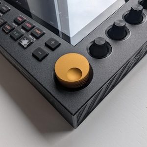 May include: Close-up of a black electronic device with a screen and various buttons and knobs. A large, gold-colored knob is prominent on the lower left. The device has a sleek, modern design, with a textured surface and a white background.