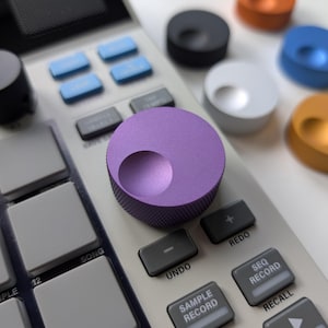 May include: A digital music production device with a purple knob and various coloured knobs. The device has grey buttons with text like "UNDO" and "SAMPLE RECORD". Blue and grey buttons are also visible.