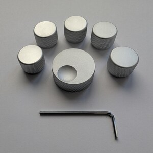 Peut inclure: Un ensemble de six boutons argent&eacute;s avec une prise textur&eacute;e, dispos&eacute;s autour d'un bouton plus grand avec une d&eacute;coupe circulaire. Une cl&eacute; Allen argent&eacute;e est &eacute;galement incluse, le tout sur une surface gris clair.
