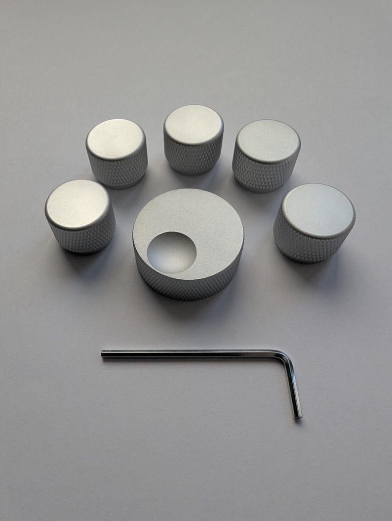 Peut inclure: Un ensemble de six boutons argent&eacute;s avec une prise textur&eacute;e, dispos&eacute;s autour d'un bouton plus grand avec une d&eacute;coupe circulaire. Une cl&eacute; Allen argent&eacute;e est &eacute;galement incluse, le tout sur une surface gris clair.
