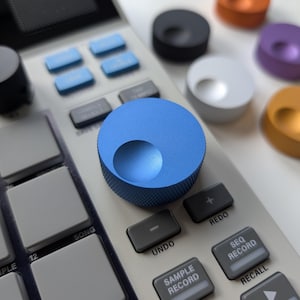 Può includere: Primo piano di un dispositivo di produzione musicale digitale con una manopola blu e vari pulsanti grigi. Manopole colorate aggiuntive in nero, arancione, bianco, viola e oro sono sparse intorno al dispositivo. I pulsanti sono etichettati "UNDO", "SAMPLE RECORD" e "SEQ RECORD".