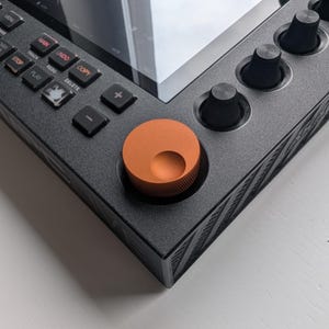 Puede incluir: Primer plano de un dispositivo negro de producción musical con un dial naranja y varias perillas negras. El dispositivo tiene una pantalla táctil y botones etiquetados como "Main", "Undo", "Copy", etc. Sobre una superficie blanca.