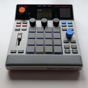 May include: An Akai MPC Sample music production controller featuring a silver chassis, a grid of sixteen grey pads, a central display screen, an orange knob, a blue knob, three black knobs, a fader, and various buttons labelled with text.
