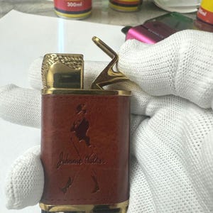 May include: A gold and brown lighter with a John Walker logo and a flame coming out of the top.